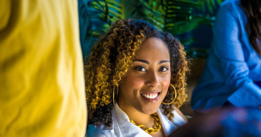 Smiling woman with shoulder-length curly hair with golden highlights, gold hoop earrings and a gold chain necklace, seated in front of tropical plants