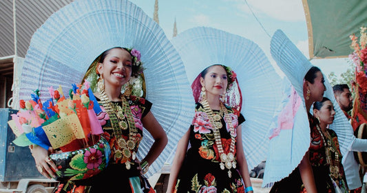 Smiling women in black huipiles with bright floral embroidery, gold coin necklaces and white pleated lace headdresses at a festival in Oaxaca de Juárez