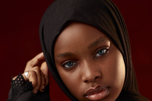 Black woman wearing a black jersey hijab, resting one hand near her temple, against a deep red backdrop