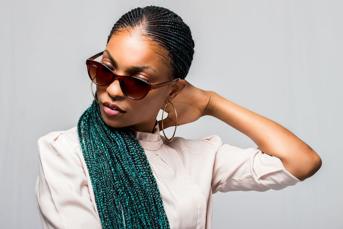 Black woman with fresh cornrows pulled back into long teal braids, resting her hand at the back of her neck