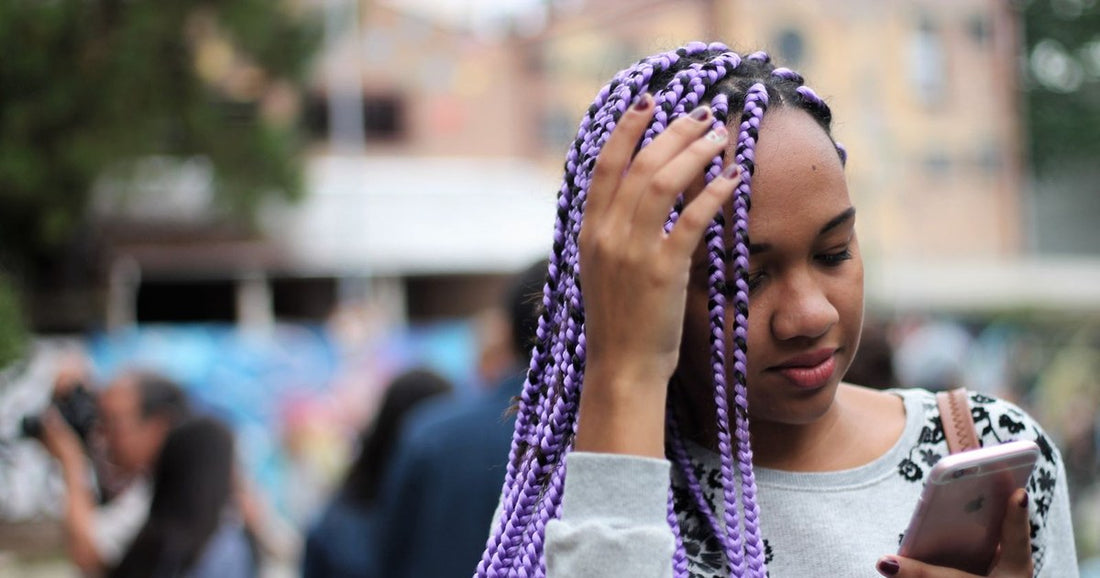 Young Black woman with long lavender box braids lifting a hand to her scalp while checking her phone outdoors