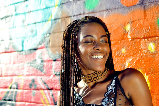 Black woman with long locs laughing in front of a colorful painted brick wall