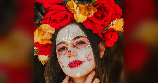 Woman in Catrina face paint with rhinestones wearing a large red and gold rose crown over long dark hair for Día de Muertos